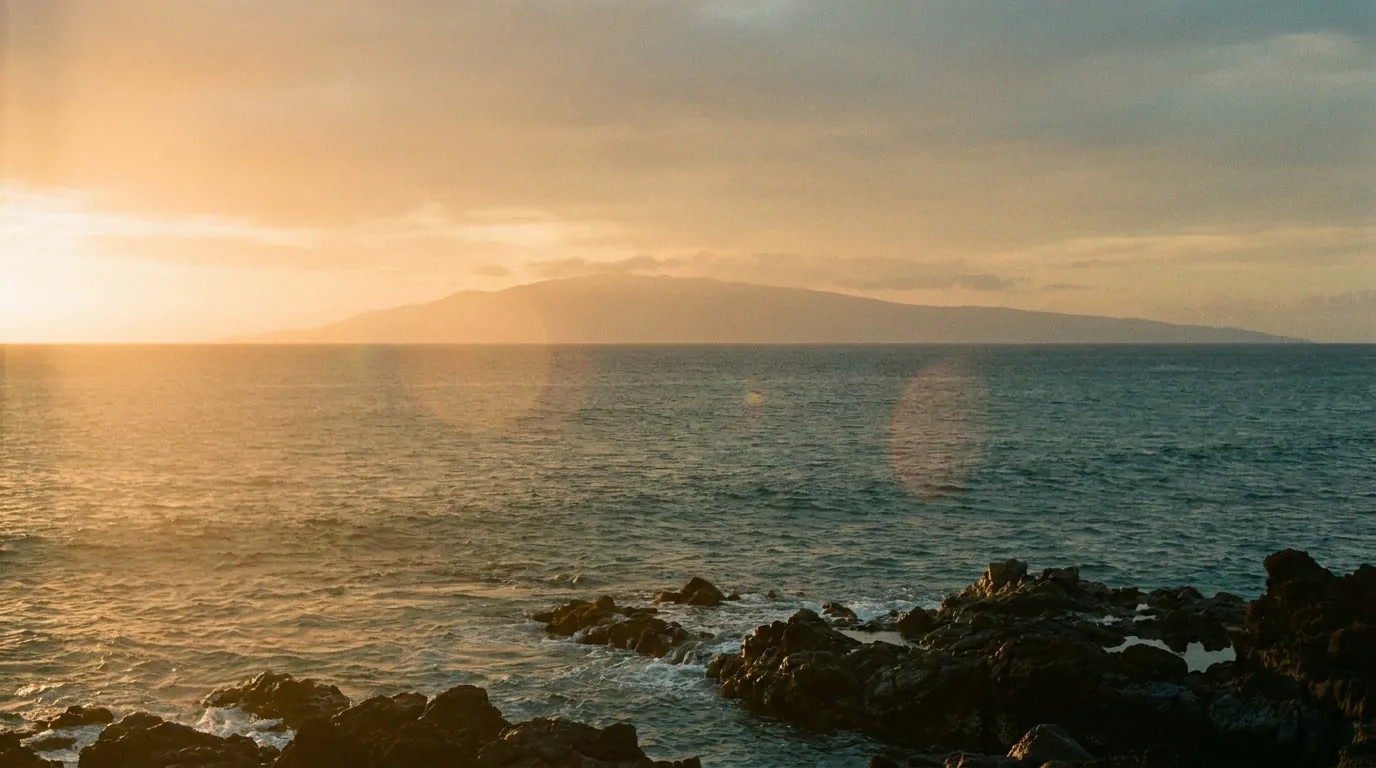 Golden-hour light over dark lava rock and calm water on Maui’s west side, an island silhouetted on the horizon